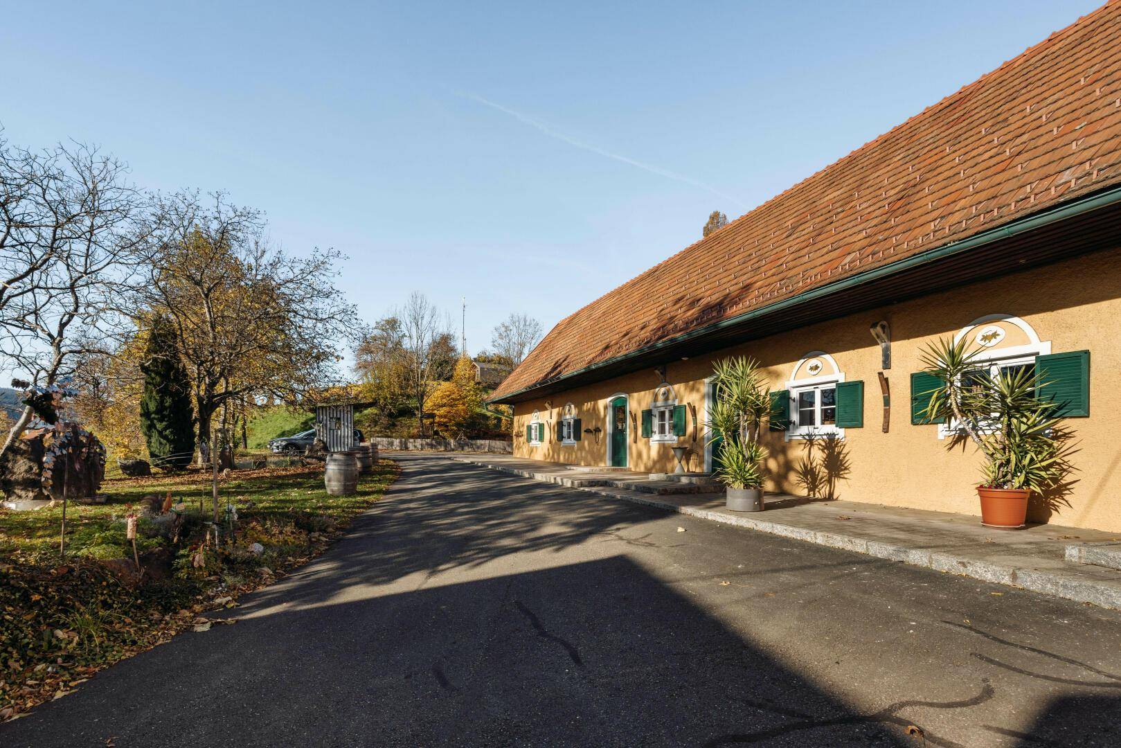 Langes gelbes Gebäude mit rotem Ziegeldach und grünen Fensterläden entlang einer Zufahrt.