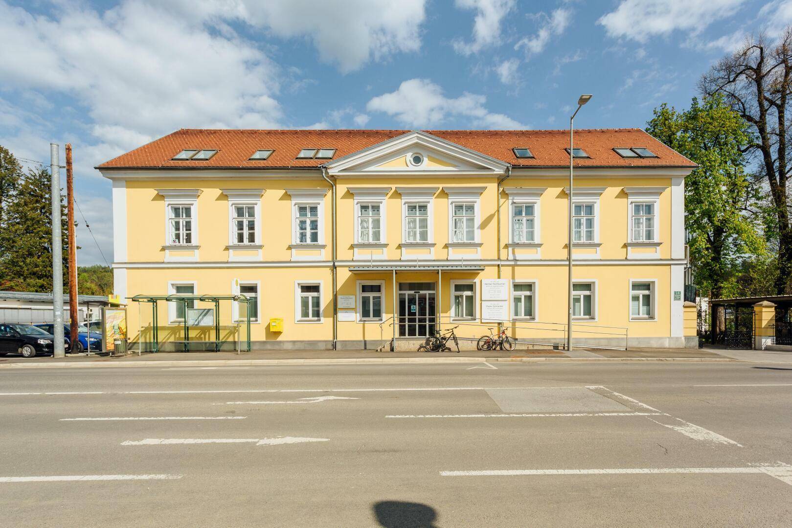 Historisches Gebäude mit gelber Fassade, vielen Fenstern und rotem Ziegeldach an der Straße.