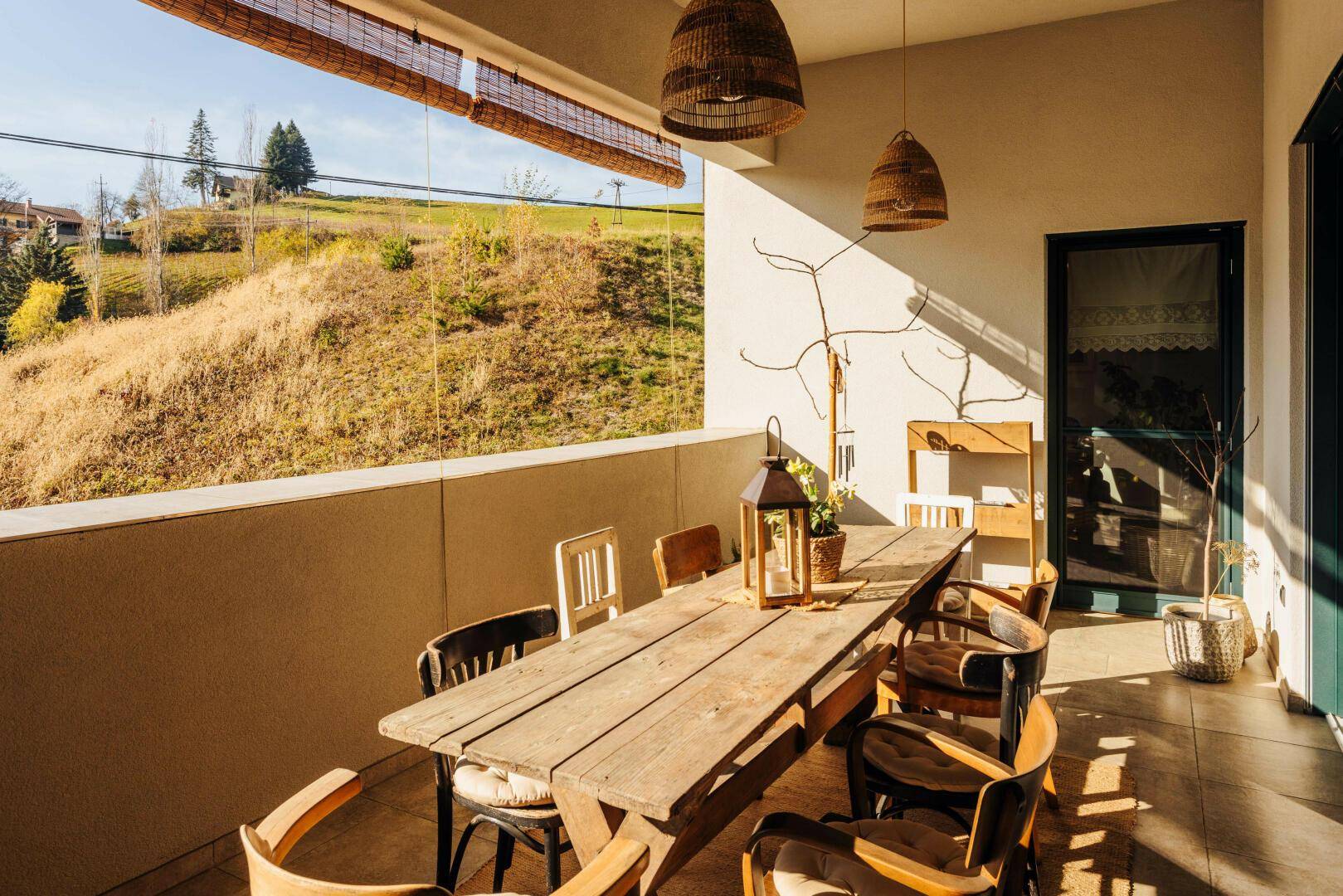 Sonniger Balkon mit großem Holztisch, Korblampen und Blick ins Grüne.