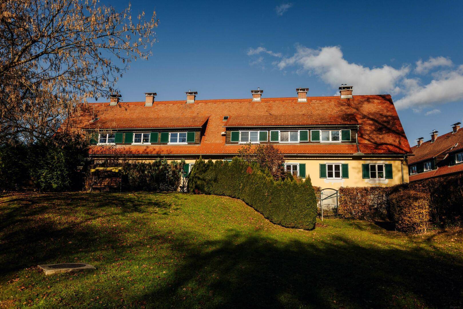 Traditionelles Wohnhaus mit gelber Fassade, roten Ziegeldächern und grünen Fensterläden, umgeben von gepflegtem Rasen.