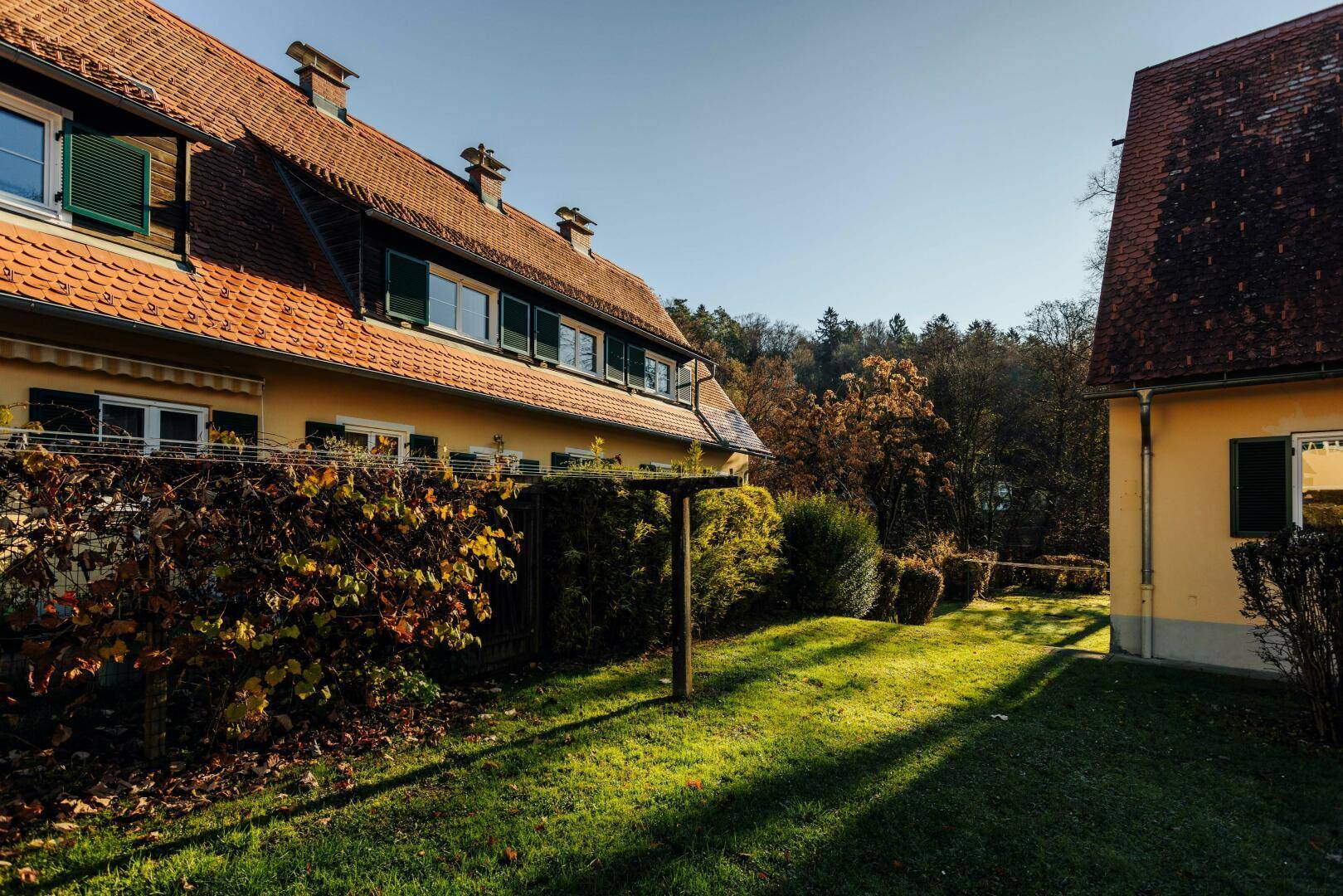 Seitenansicht eines Wohnhauses mit gelber Fassade, roten Ziegeldächern und grünen Fensterläden, umgeben von Natur.