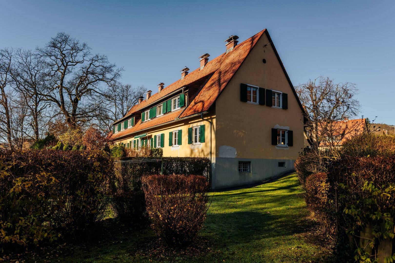 Traditionelles Haus mit rotem Ziegeldach und grünen Fensterläden, eingebettet in eine grüne Hanglage.