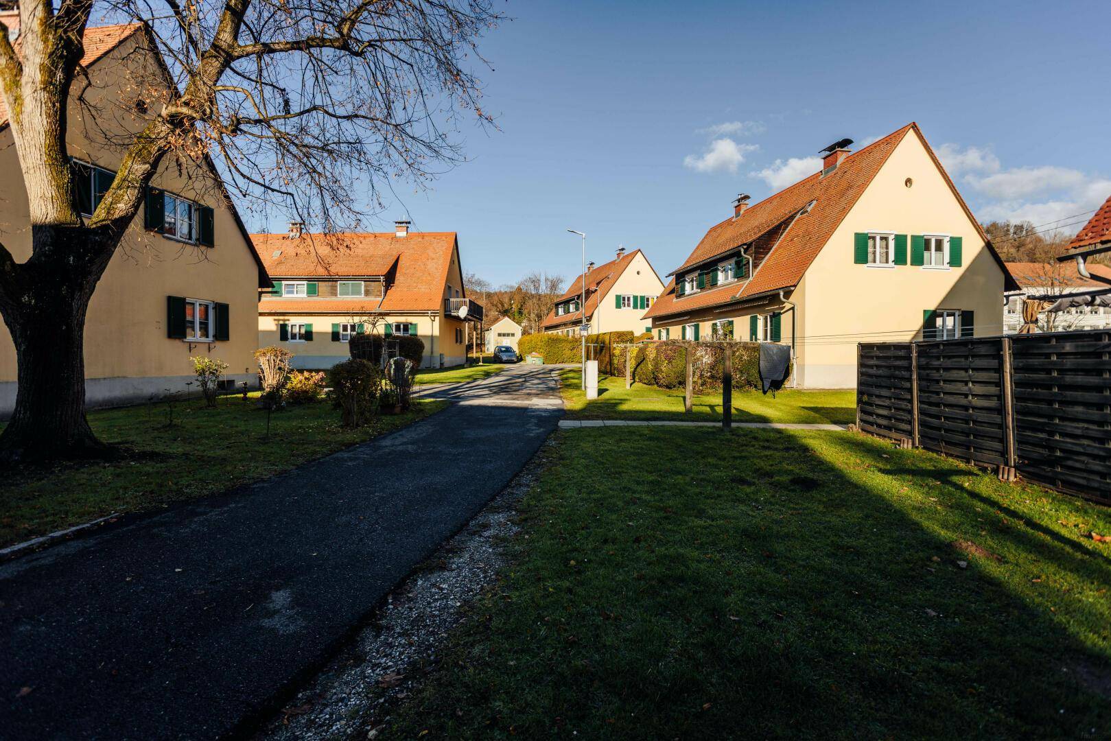Eine ruhige Wohnstraße mit traditionellen Häusern, umgeben von Grünflächen und Bäumen unter blauem Himmel.