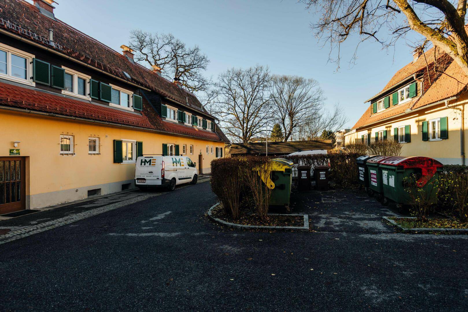Eine Wohnstraße mit traditionellen Häusern und Mülltonnen, umgeben von Bäumen und Sträuchern.