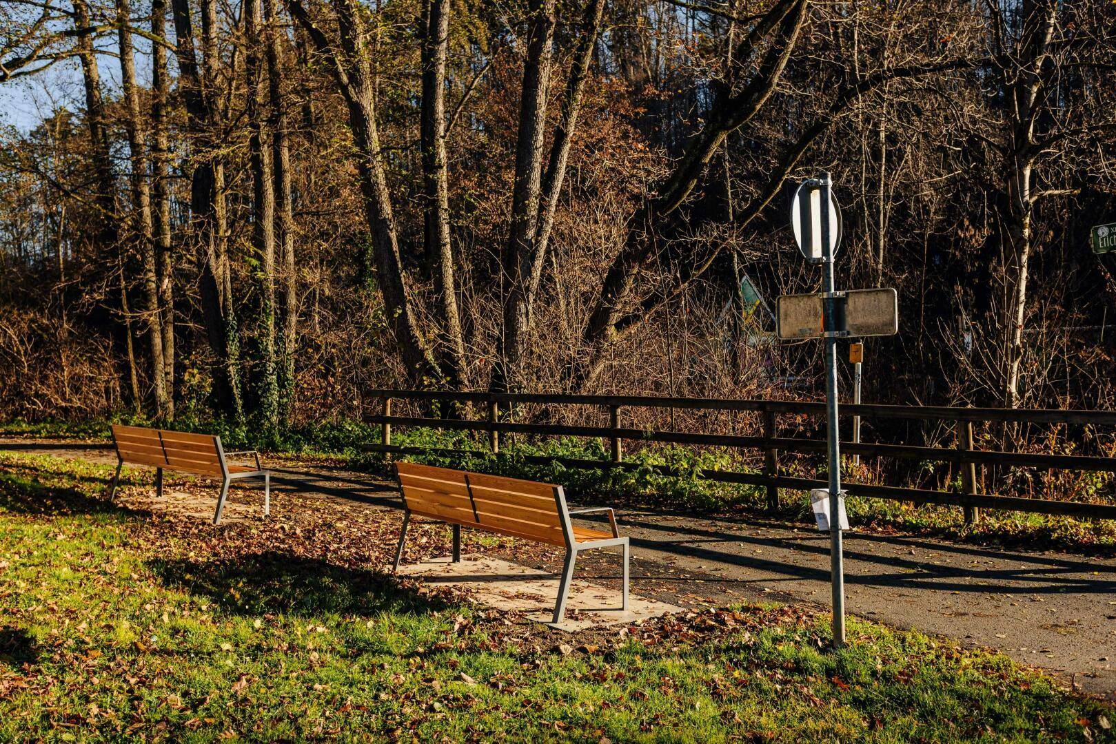 Ein sonniger Weg am Waldrand mit Holzbänken und einem Verkehrsschild, umgeben von Bäumen.