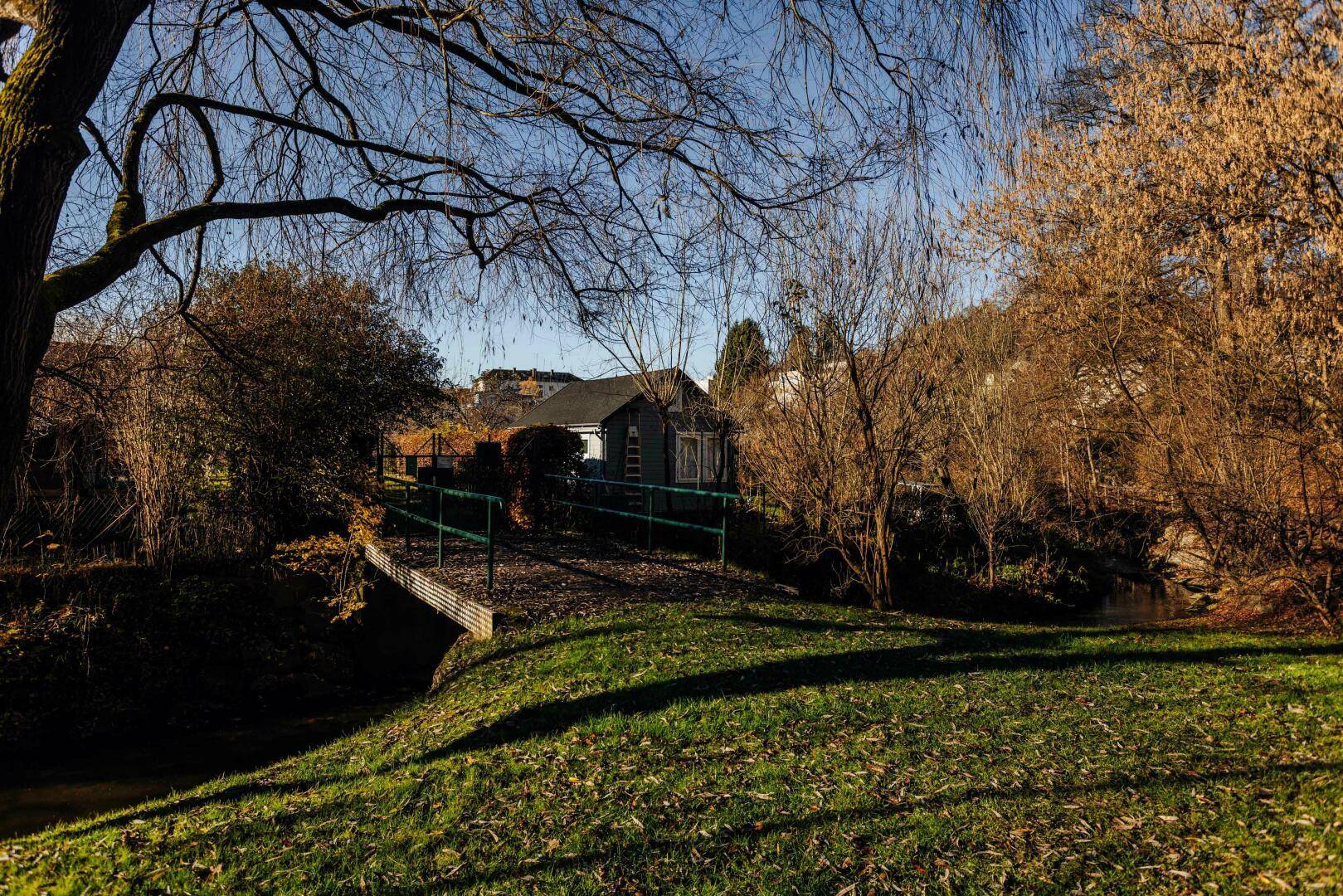 Ein kleines Haus über eine Brücke in einer idyllischen, grünen Umgebung mit Bäumen und einem Bach.