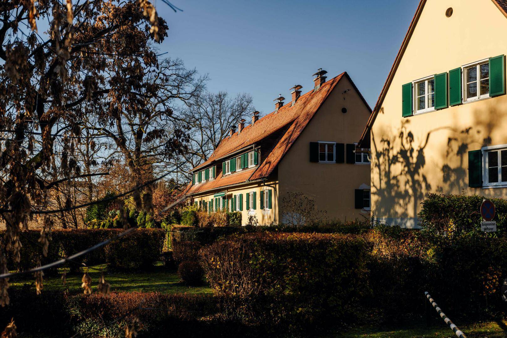 Traditionelle Häuser mit roten Ziegeldächern und grünen Fensterläden, umgeben von gepflegten Hecken und Bäumen.