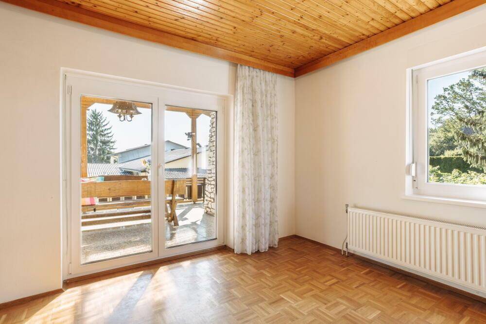 Heller Raum mit Holzbalkendecke, Parkettboden und Zugang zu einem sonnigen Balkon.