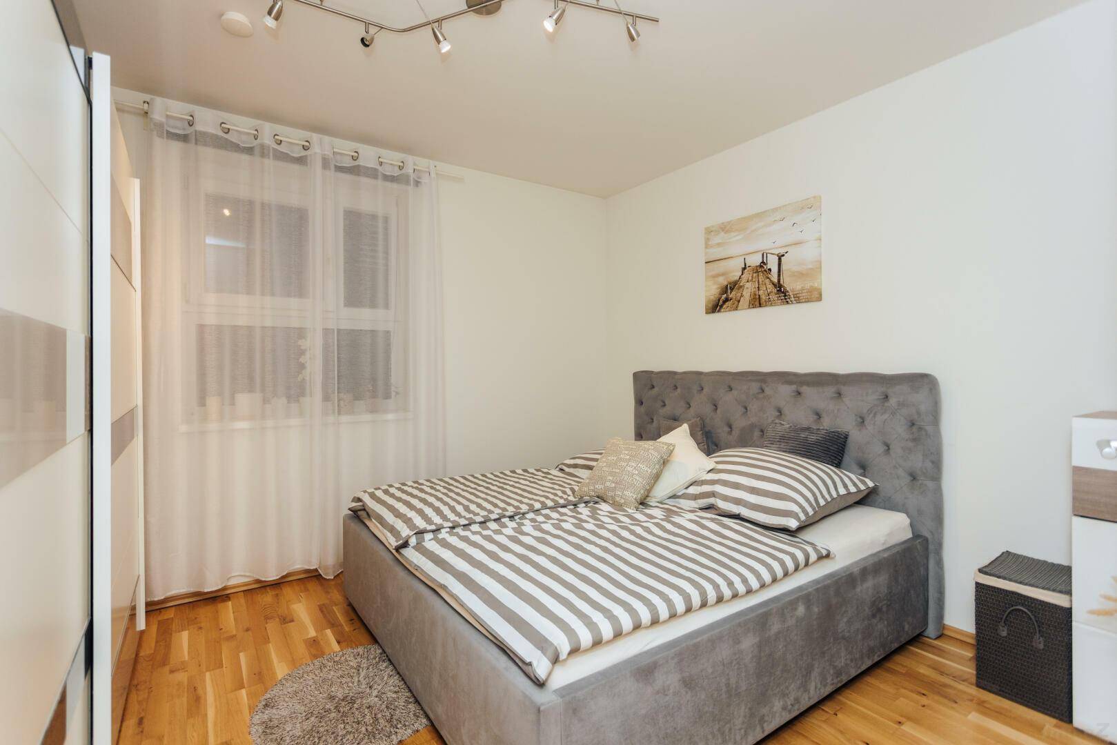 Helles Schlafzimmer mit modernem Polsterbett, Holzfußboden und großem Fenster mit Vorhängen.