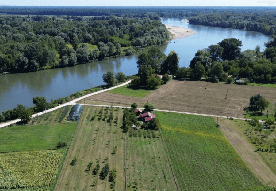 Luftaufnahme eines Anwesens mit Obstbäumen, Feldern und einem Fluss im Hintergrund unter blauem Himmel.