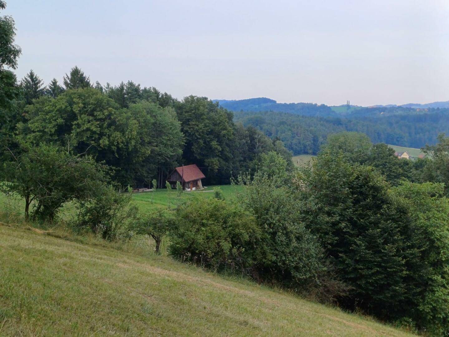 Panoramablick auf die kleine Holzhütte in einer weiten, hügeligen Landschaft mit viel Grün.
