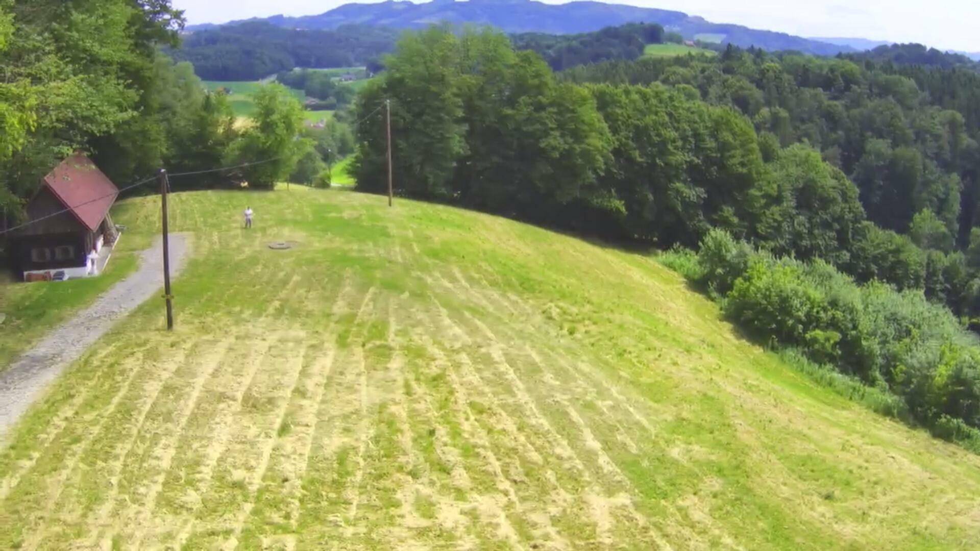Luftaufnahme der Holzhütte und der weitläufigen, gemähten Wiesen, umgeben von bewaldeten Hügeln.