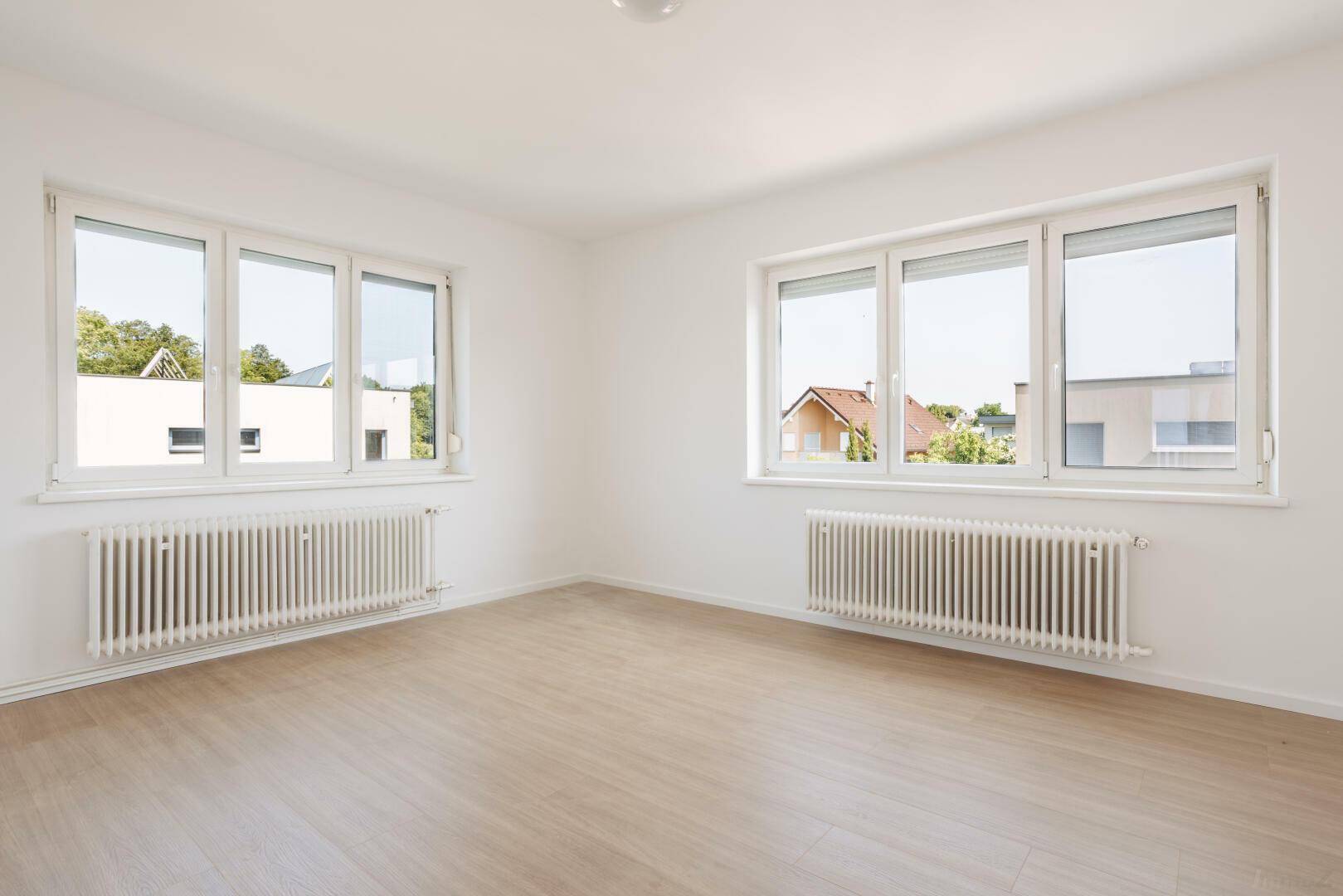 Geräumiges Wohnzimmer mit zwei großen Fenstern und hellem Laminatboden.