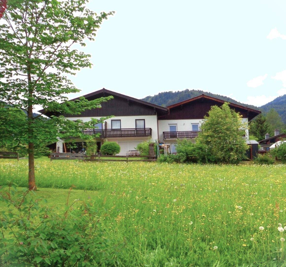Hausansicht mit einem blühenden Feld im Vordergrund, umgeben von Bäumen und Bergen.