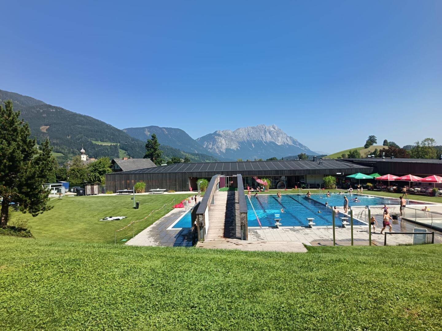 Belebter Außenpoolbereich mit Badegästen, umgeben von grünen Wiesen und einer Berglandschaft.