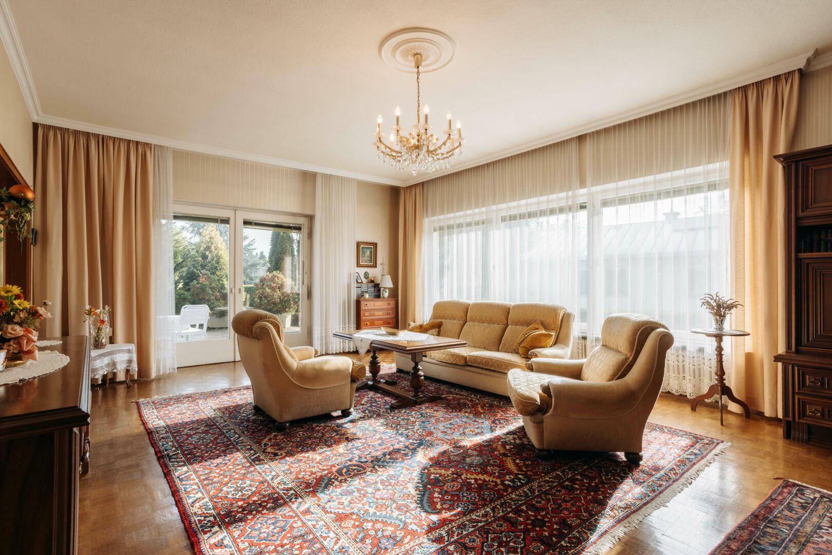 Geräumiges Wohnzimmer mit traditionellen Möbeln, großem Teppich und viel natürlichem Licht durch die Fenster.