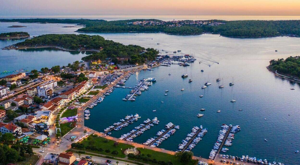 Luftaufnahme einer Küstenstadt mit belebtem Yachthafen bei Sonnenuntergang, umgeben von Inseln und Meer.
