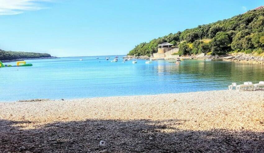 Kiesstrand mit kristallklarem Wasser, umgeben von grünen Hügeln und Booten in der Bucht.