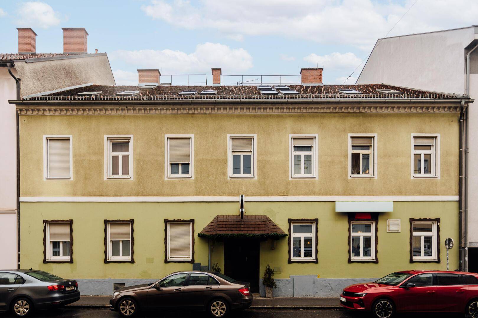 Historisches Gebäude mit beiger und hellgrüner Fassade, vielen Fenstern mit Rollläden und parkenden Autos davor an einer Straße.
