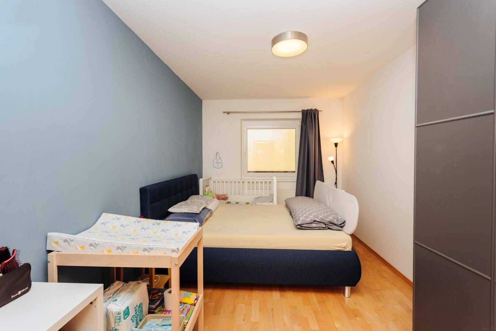 Schlafzimmer mit einem Bett, Babybett und einer blauen Akzentwand, ausgestattet mit Holzboden.