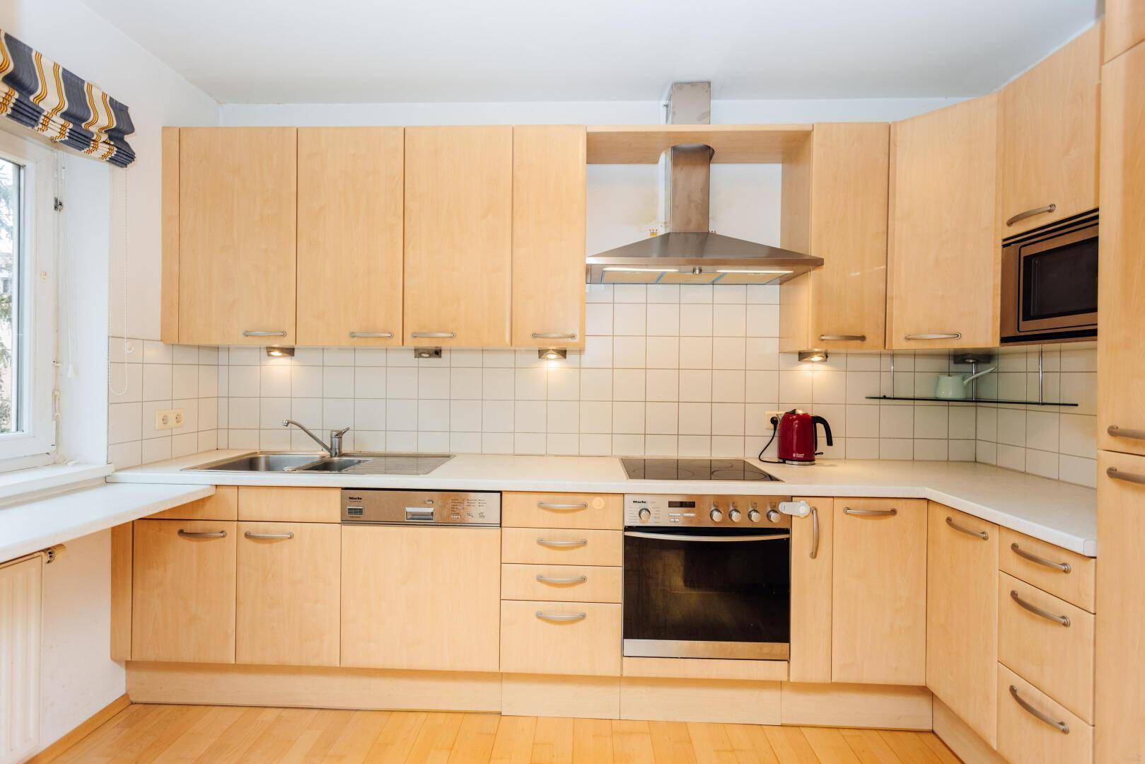 Einbauküche mit hellen Holzschränken, modernen Geräten und einem Fenster für Tageslicht.