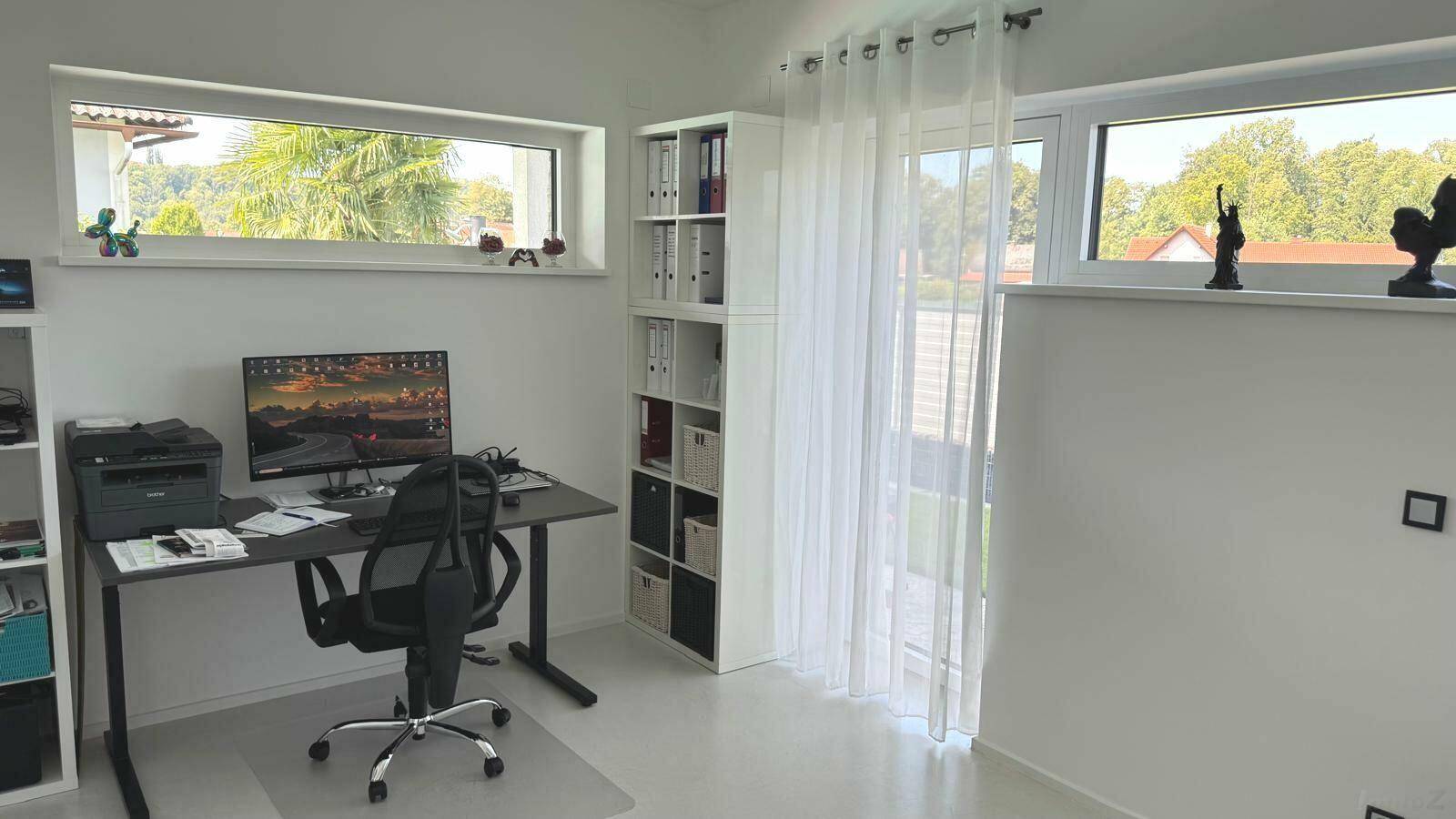 Helles Arbeitszimmer mit Schreibtisch, Regalen und großen Fenstern mit Blick ins Grüne.