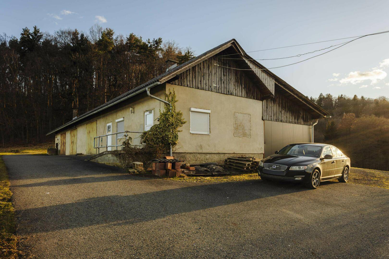 Langes landwirtschaftliches Gebäude mit Parkplatz davor, umgeben von ländlicher Umgebung.