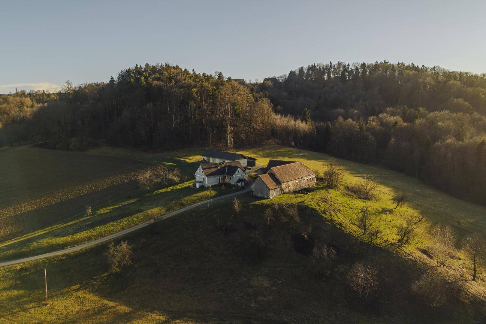 Luftaufnahme eines ländlichen Anwesens mit mehreren Gebäuden, eingebettet in grüne Hügel und Wald.