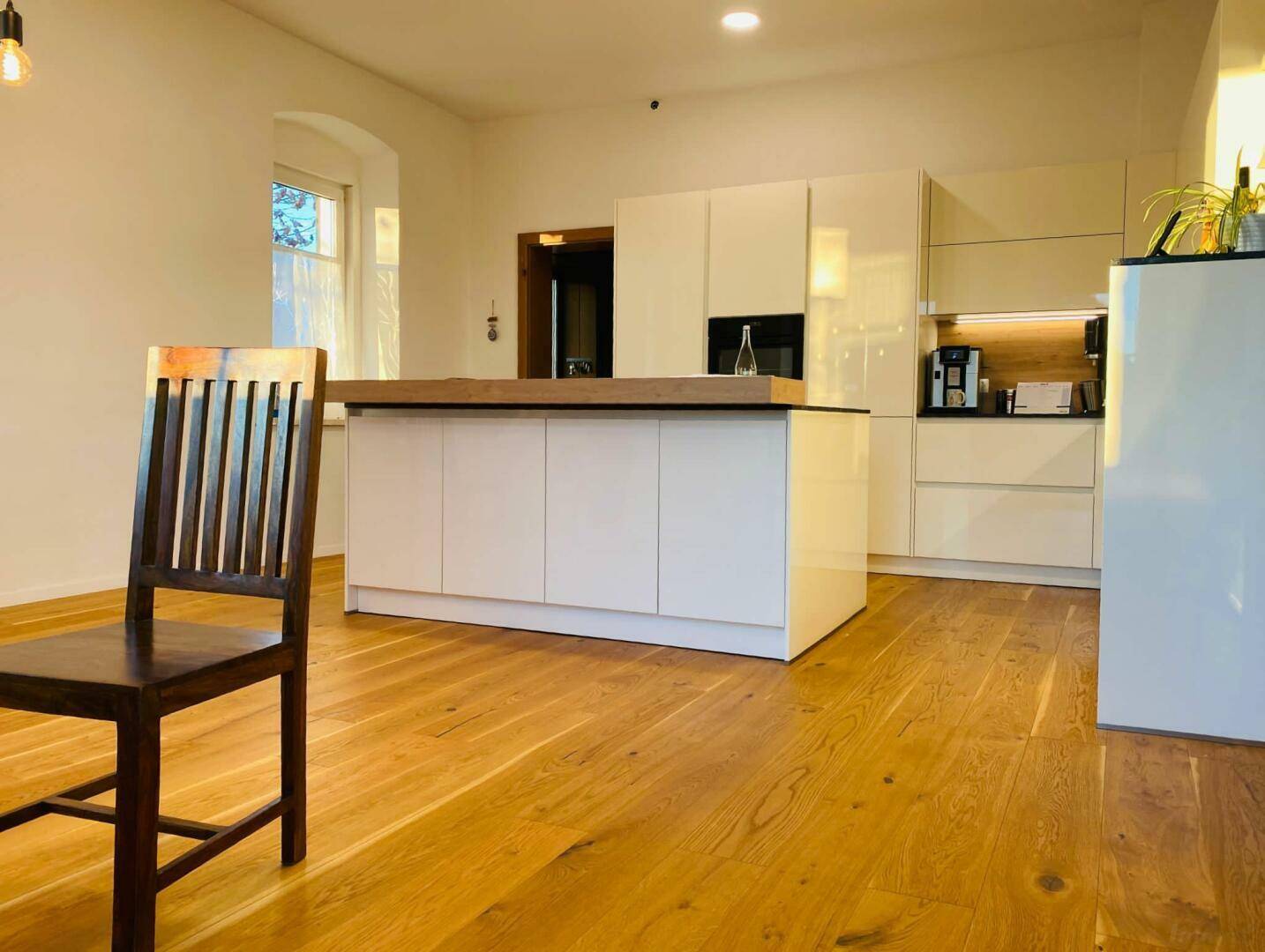 Moderne weiße Küche mit Kücheninsel, integrierten Geräten und Holzboden in hellem Raum.