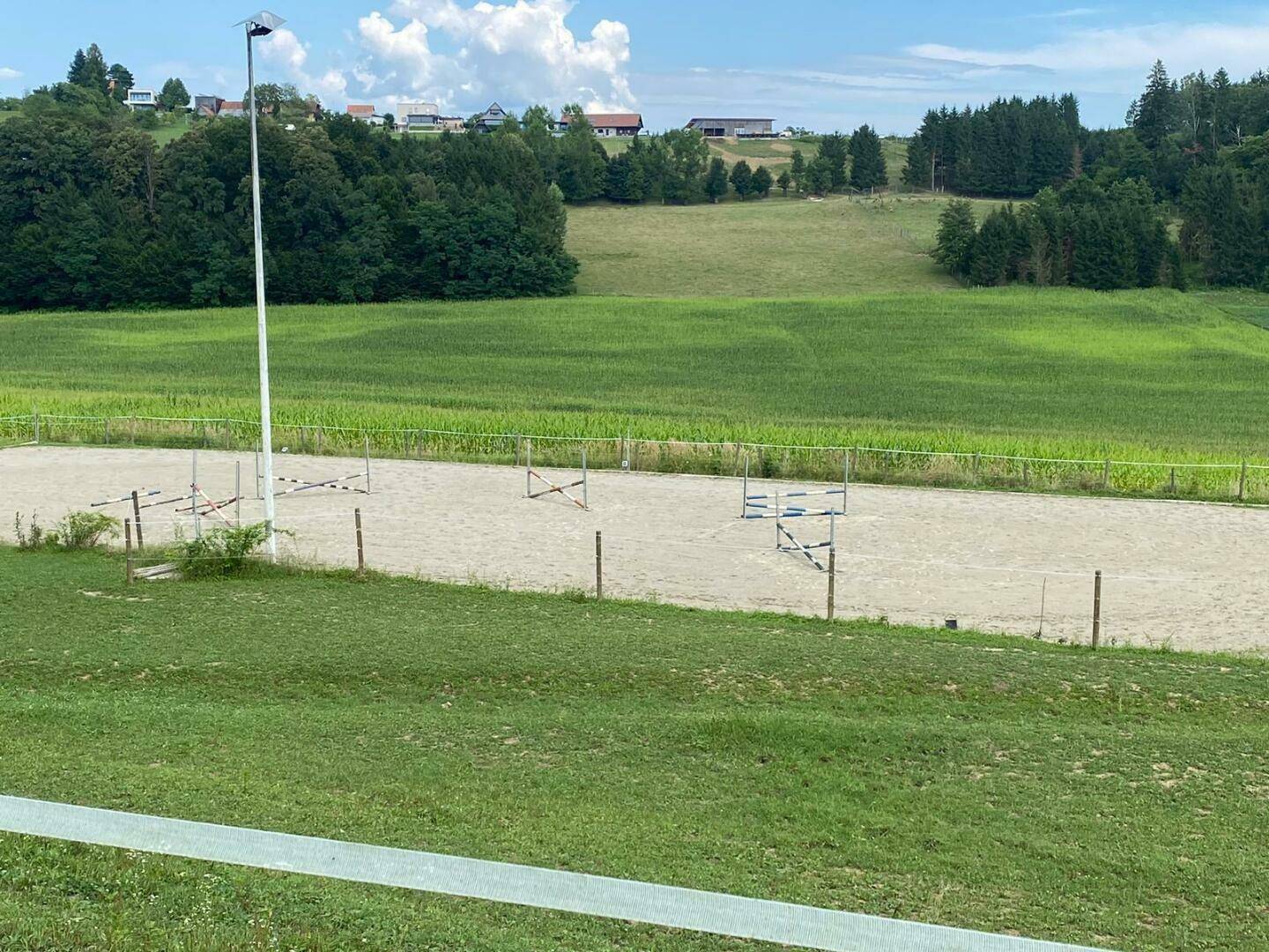 Weitläufiger Reitplatz in ländlicher Umgebung, umgeben von grünen Wiesen und Wäldern unter blauem Himmel.