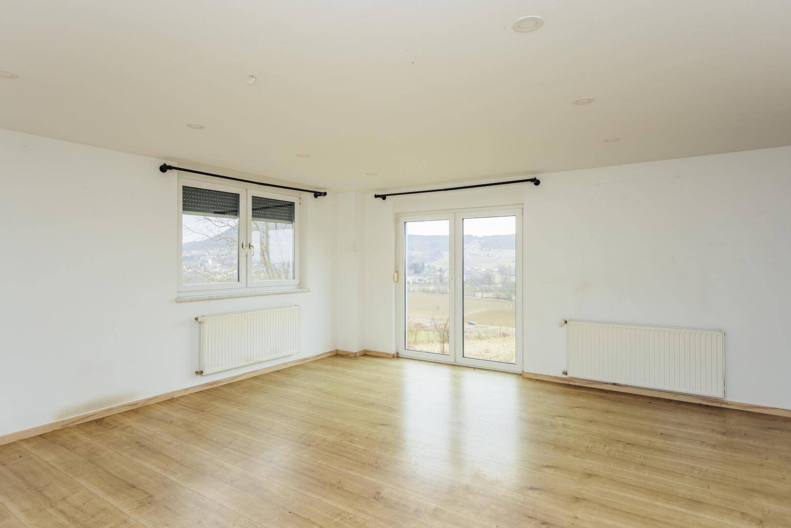 Heller, leerer Raum mit Holzboden und großen Fenstern mit Blick ins Grüne.