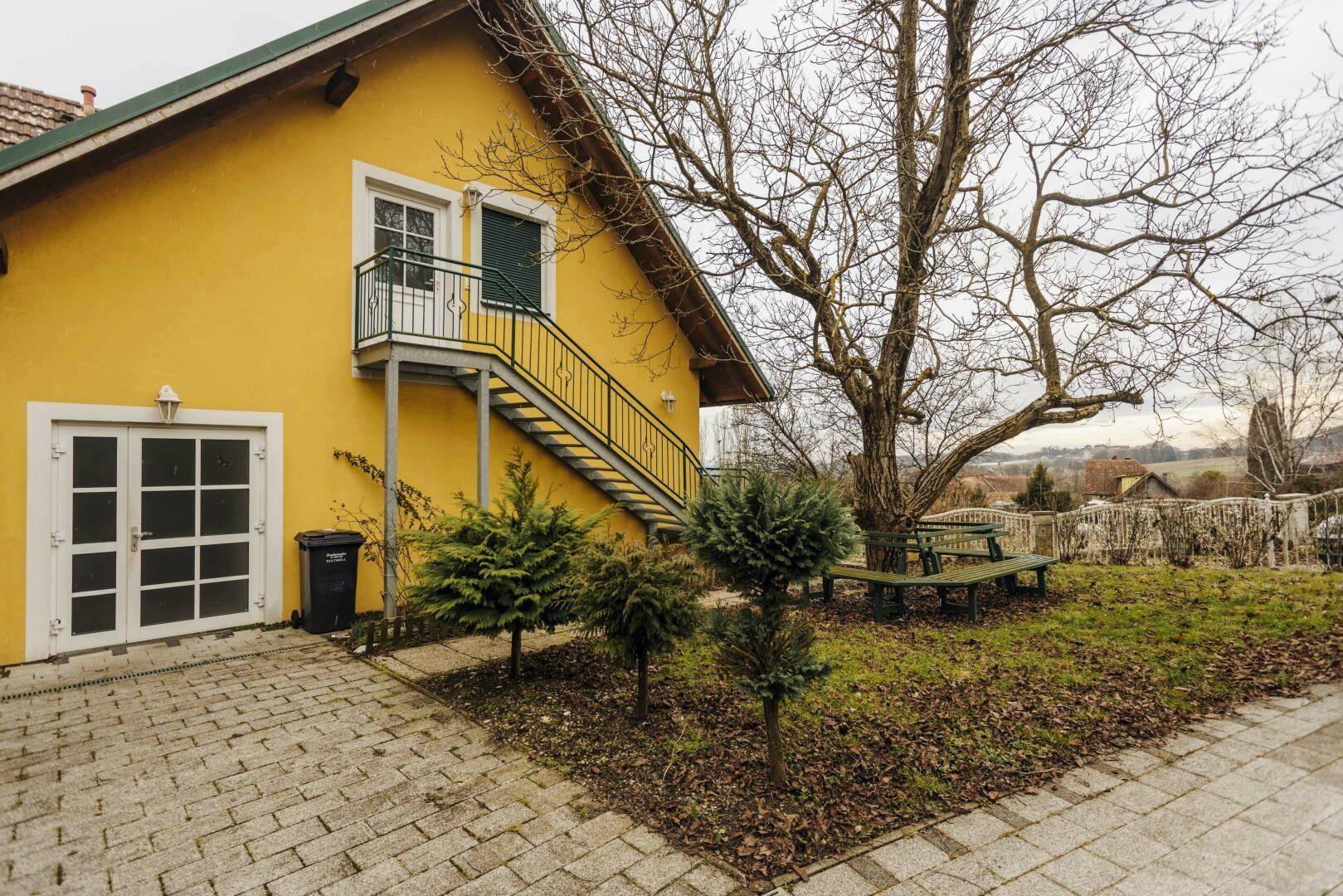 Seitenansicht des gelben Gebäudes mit Außentreppe und gepflastertem Vorplatz.