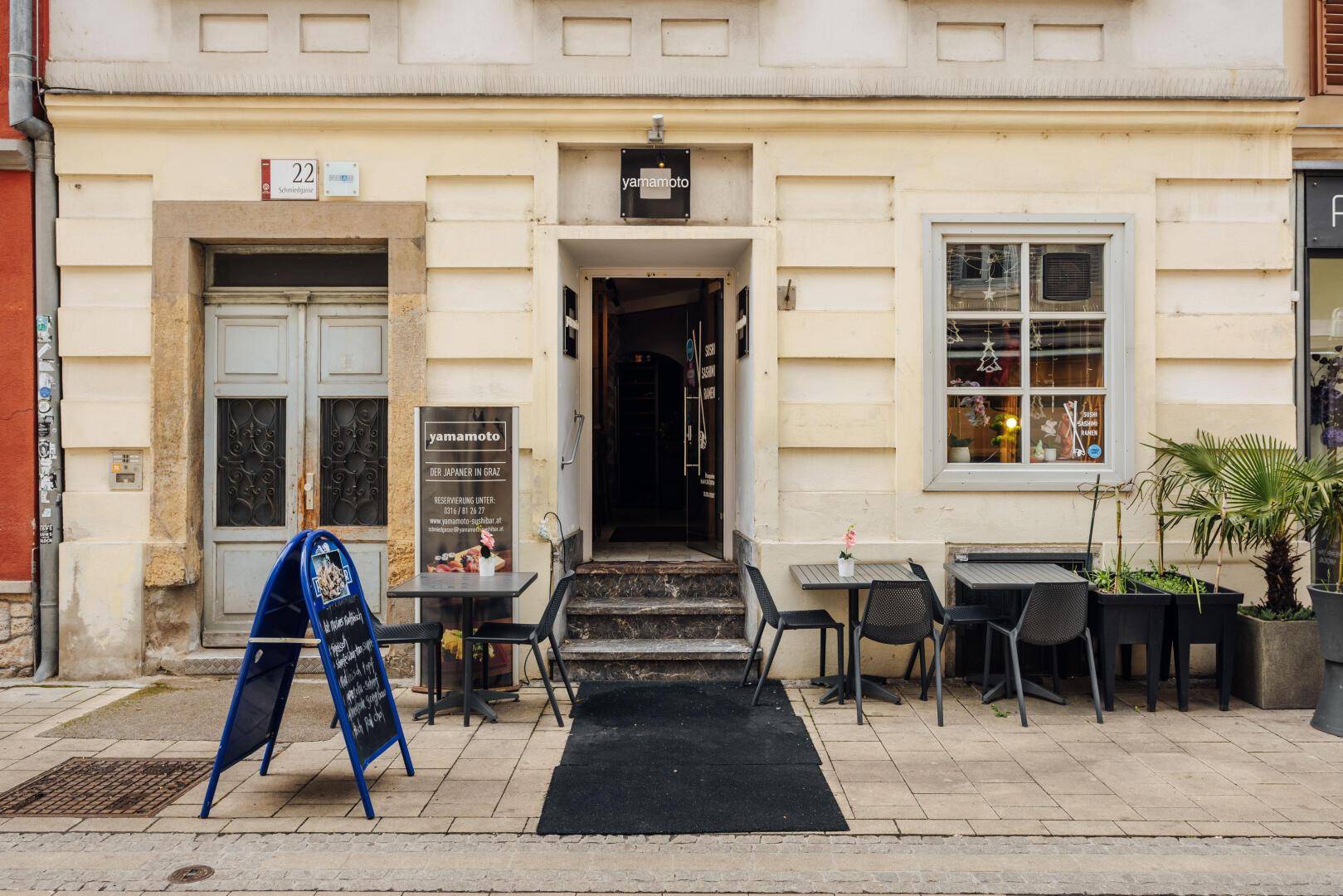 Fassade eines Restaurants mit Eingang, Außenbestuhlung und Werbetafel auf dem Bürgersteig in einer belebten Straße.