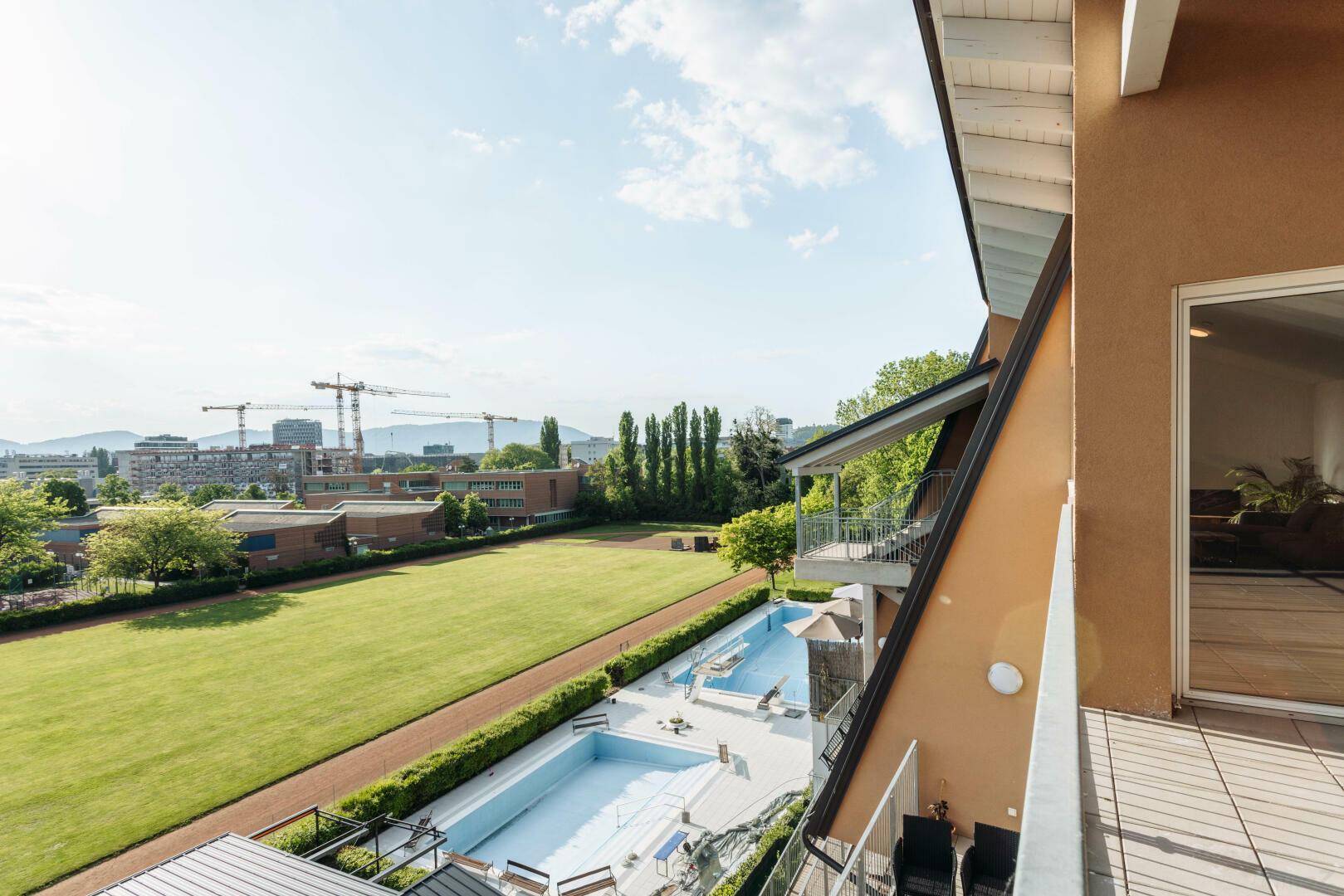 Terrasse mit weitem Blick auf einen Sportplatz, Bäume und einen großen Außenpool.