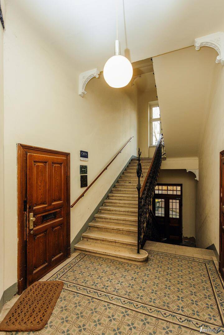 Historisches Treppenhaus mit gemusterten Fliesen, Holzgeländer und hohen Decken, zeugt von alter Baukunst.