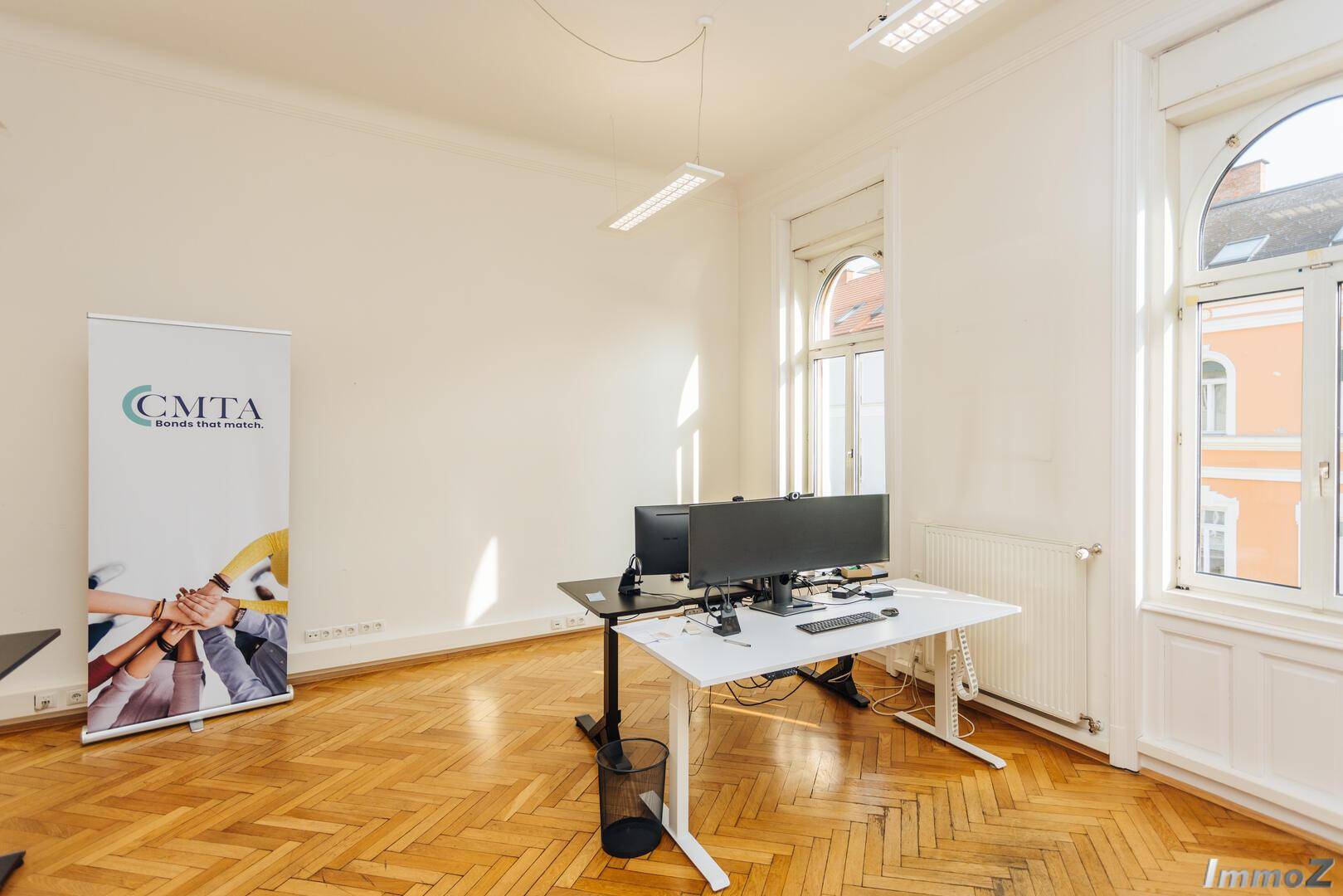 Helles Büro mit Fischgrätparkett, hohen Fenstern und einem modernen Arbeitsplatz, ideal für produktives Arbeiten.
