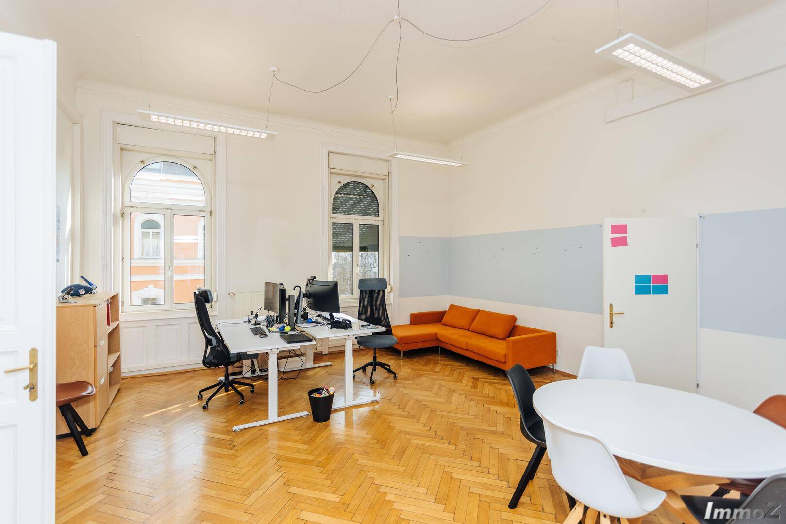 Geräumiges Büro mit Fischgrätparkett, einer orangefarbenen Couch und modernen Arbeitsplätzen, schafft eine lebendige Atmosphäre.