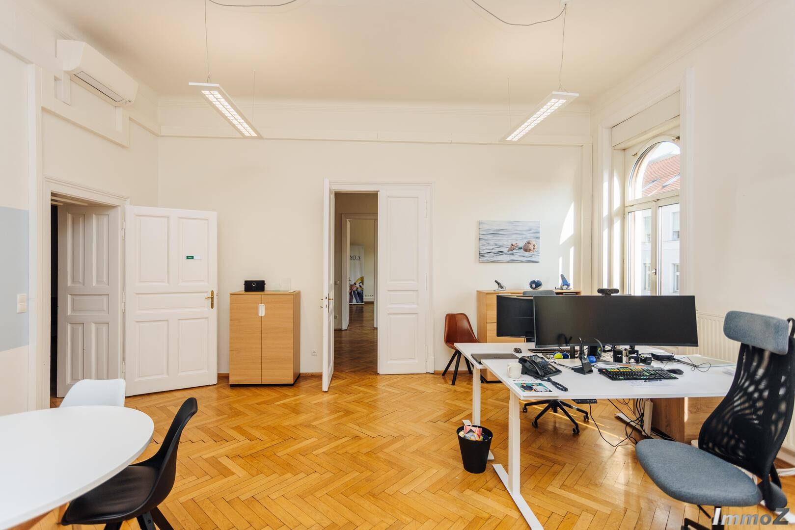 Helles Büro mit Fischgrätparkett, hohen Fenstern und modernen Arbeitsplätzen, bietet eine angenehme Arbeitsumgebung.