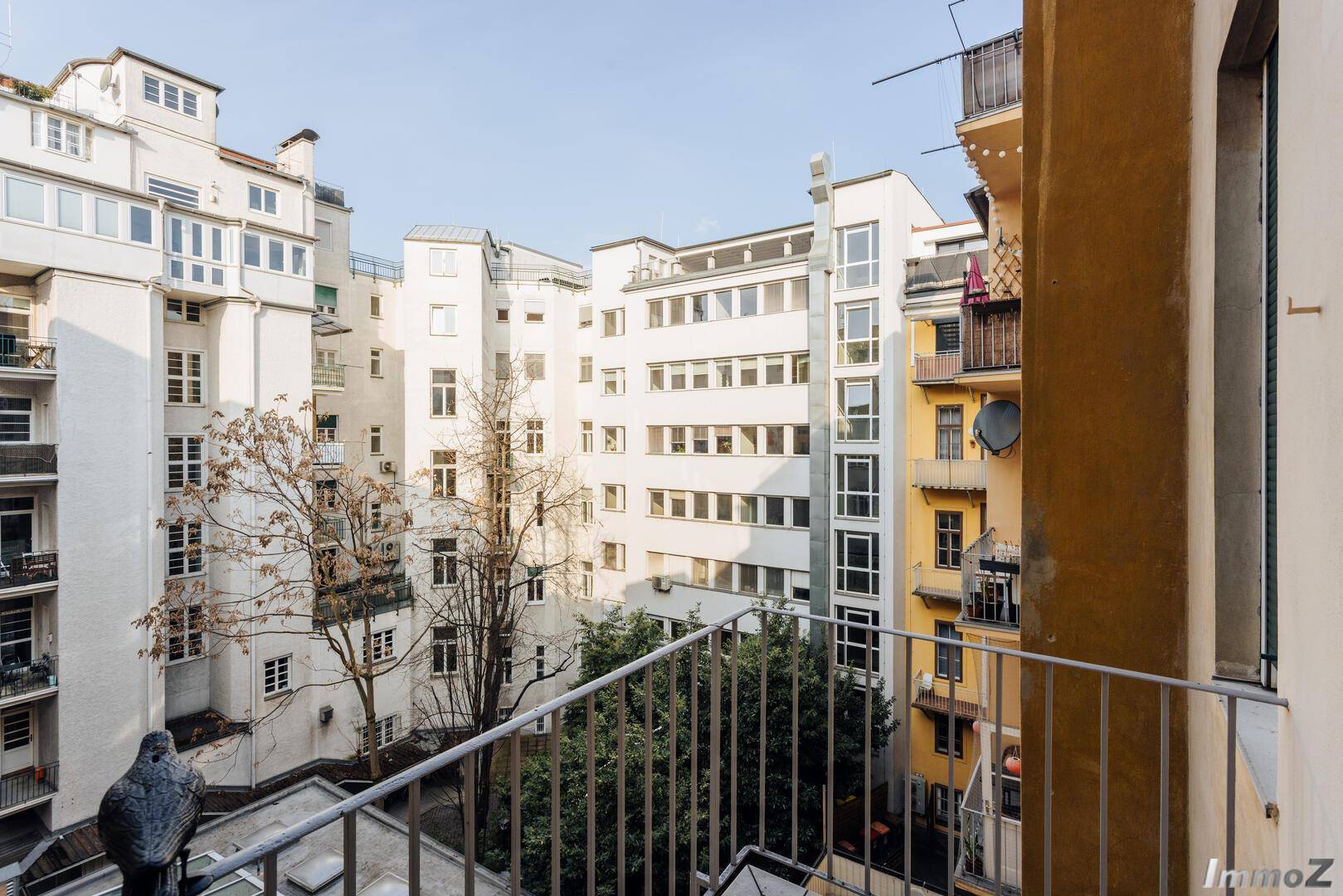 Blick vom Balkon auf einen ruhigen Innenhof mit mehreren Gebäuden und Bäumen, bietet eine grüne Oase.