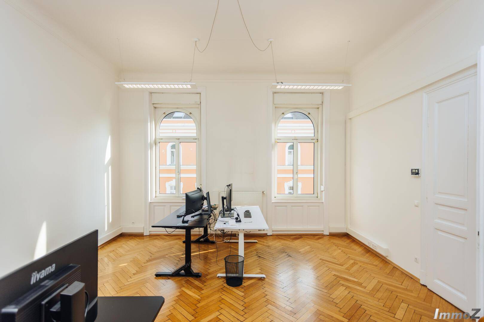 Geräumiges Büro mit Fischgrätparkett, hohen Fenstern und zwei modernen Arbeitsplätzen, bietet viel Platz.