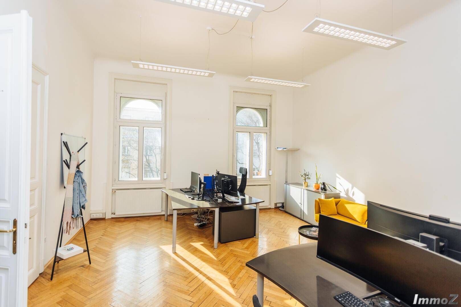 Helles Büro mit großen Fenstern, Fischgrätparkett und modernen Schreibtischen mit Computern, ideal für konzentriertes Arbeiten.