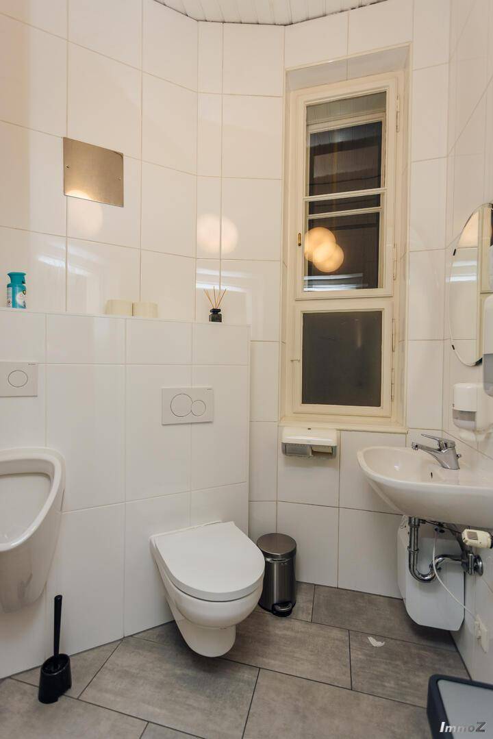 Modernes Badezimmer mit weißen Fliesen, Urinal, wandhängendem WC und einem Fenster, sorgt für gute Belüftung.