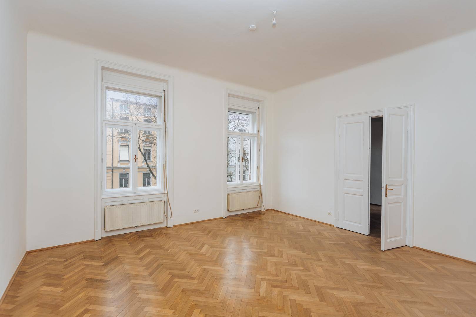 Heller Wohnraum mit klassischem Fischgrätparkett und hohen Fenstern, die den Altbaucharakter betonen.
