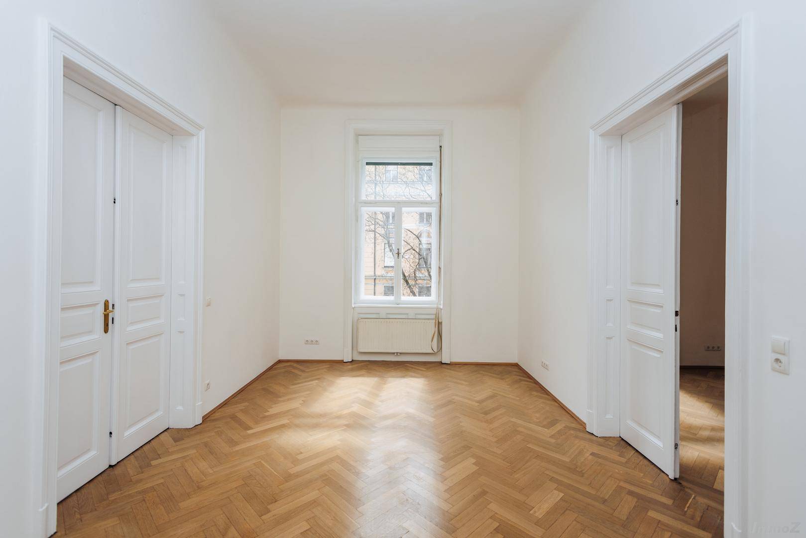 Langer, heller Raum mit Fischgrätparkett und einem Fenster, flankiert von zwei hohen Türen.