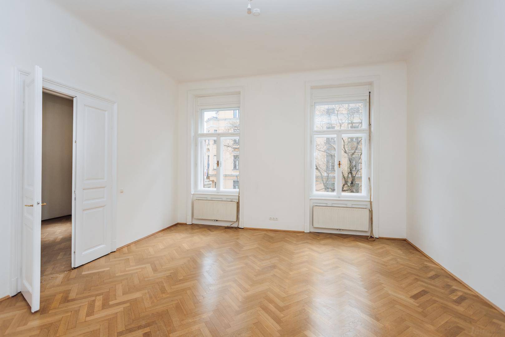 Leerer Raum mit Fischgrätparkett und zwei großen Fenstern, die den Blick auf die Umgebung freigeben.