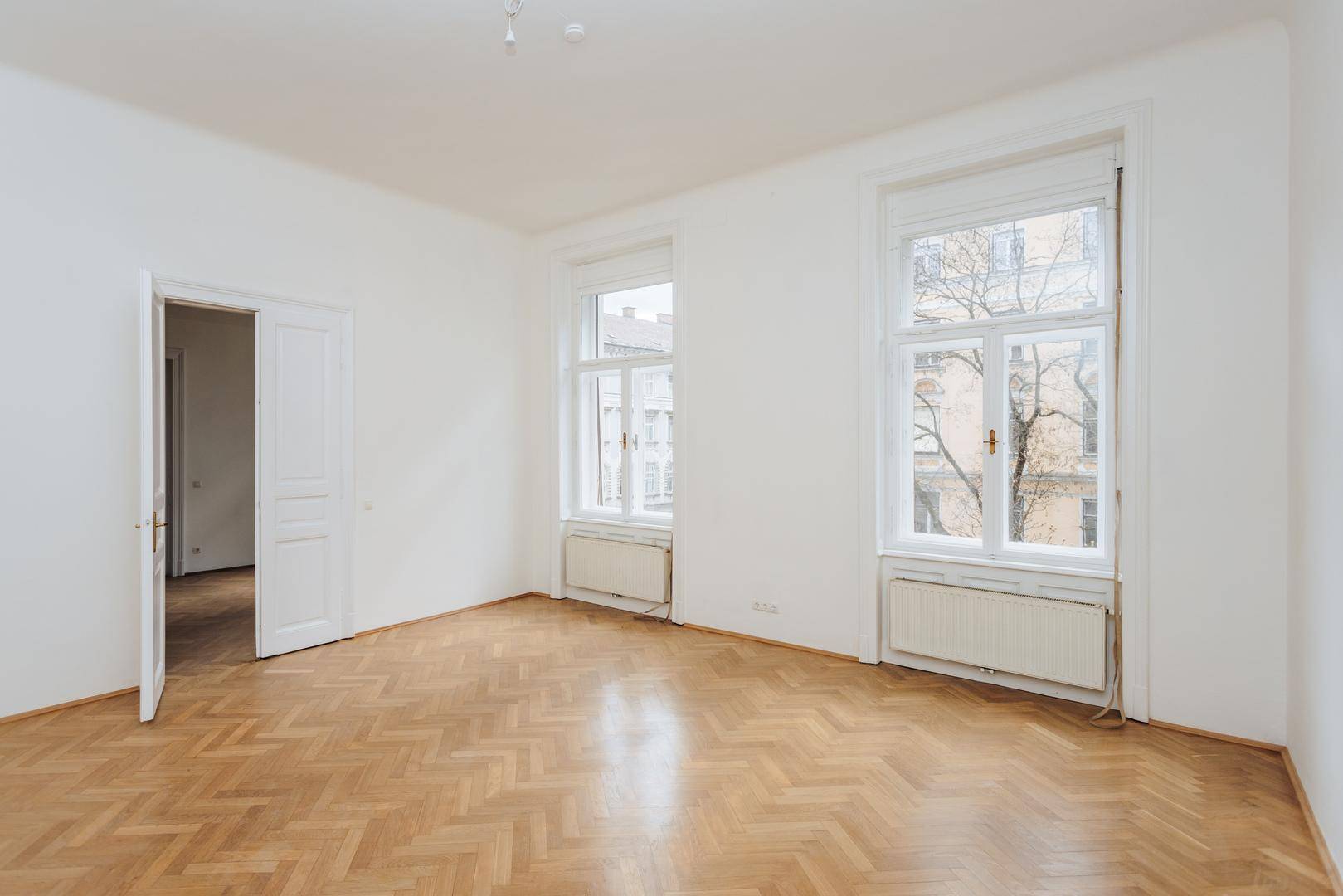Unmöblierter Wohnraum mit hellem Fischgrätparkett und zwei hohen Fenstern, die für viel Helligkeit sorgen.