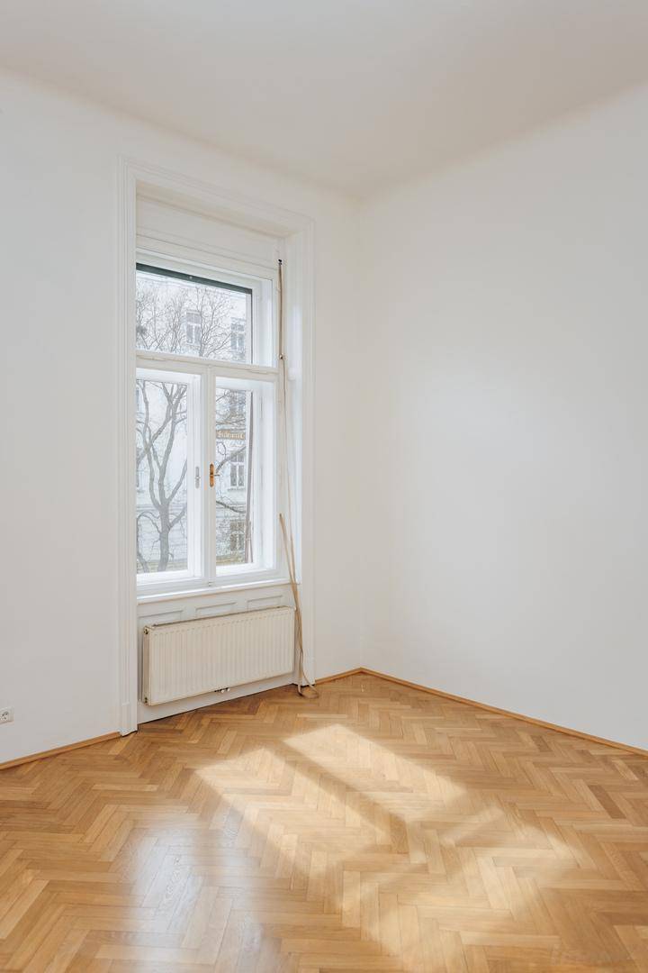 Heller Raum mit Fischgrätparkett und einem hohen Fenster, durch das Sonnenlicht einfällt.