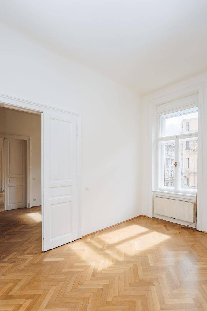 Unmöblierter Raum mit Fischgrätparkett und Sonnenlicht, das durch das Fenster auf den Boden fällt.
