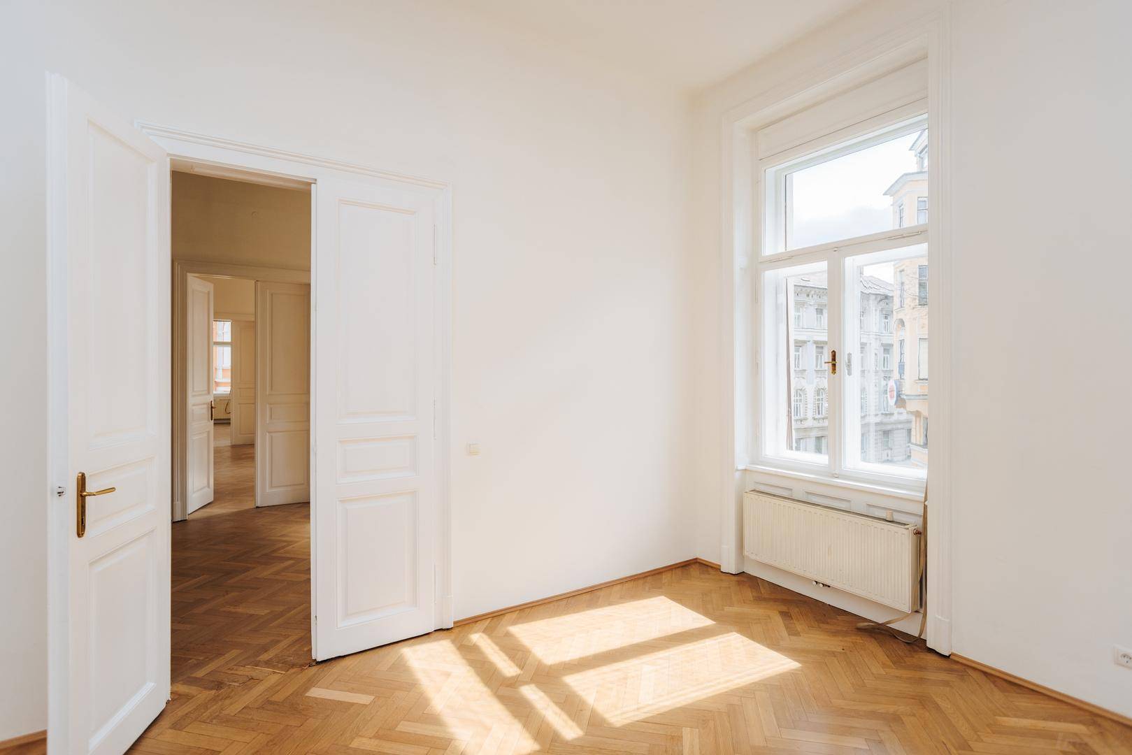 Heller Raum mit Fischgrätparkett und Sonnenlicht, das durch das Fenster auf den Boden fällt.