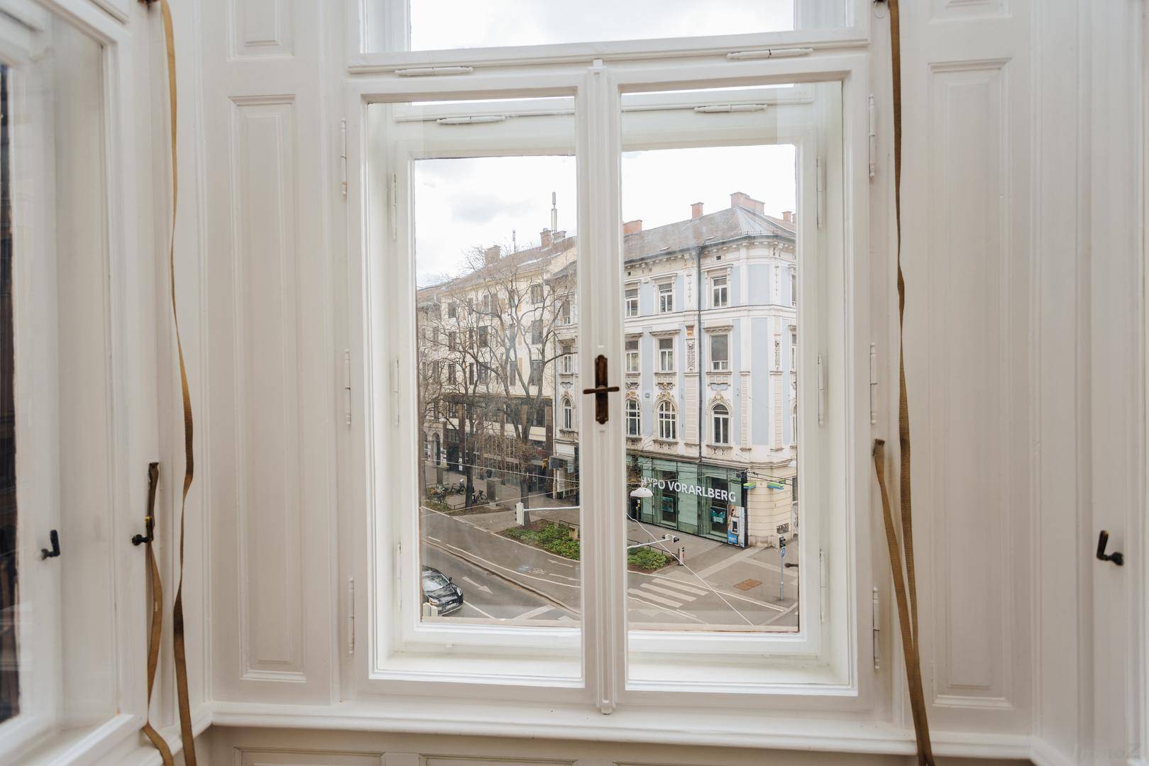 Fensterblick auf die belebte Stadtstraße und gegenüberliegende Gebäude, mit klassischem Fensterrahmen.